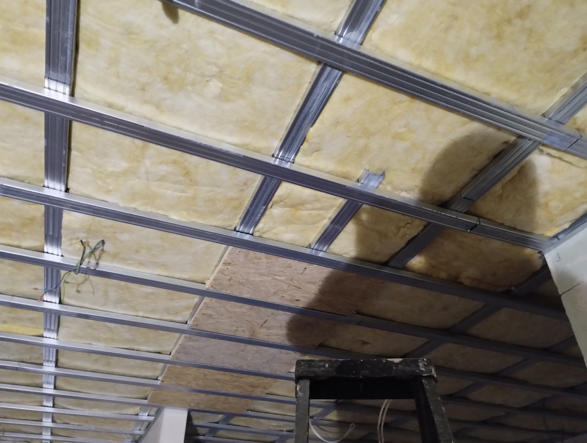 Sufit podwieszany w trakcie montażu: metalowa konstrukcja, izolacja z wełny mineralnej, fragment płyty OSB. Widoczne przewody elektryczne i drabina.