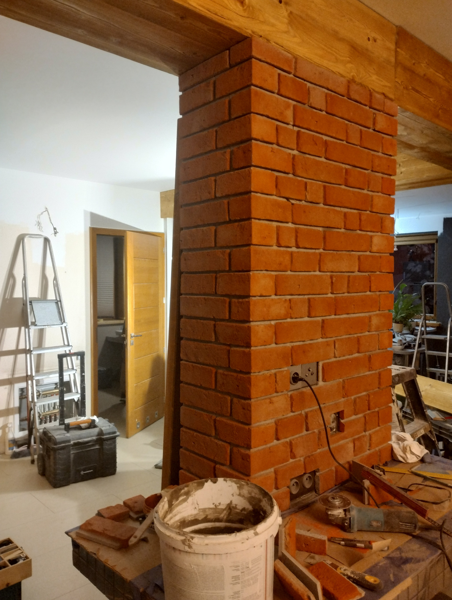 Ceglana kolumna w surowym wnętrzu z drewnianymi belkami, widoczne narzędzia i drabina, gniazdka elektryczne w ścianie, etap prac wykończeniowych.