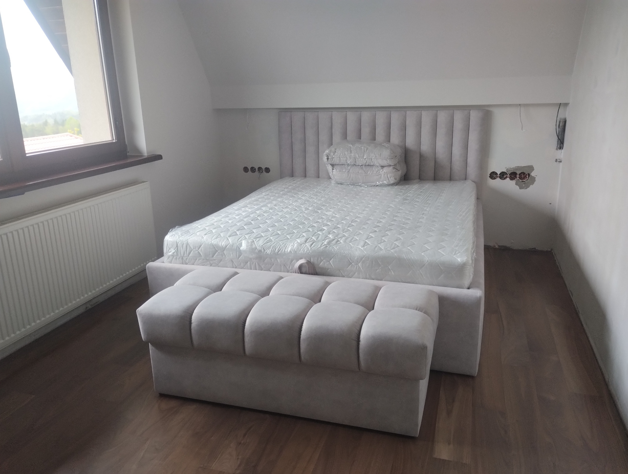 Jasne, tapicerowane łóżko z wezgłowiem i pufą na tle pokoju w trakcie wykańczania, widoczne niedokończone gniazdka elektryczne i grzejnik pod oknem.