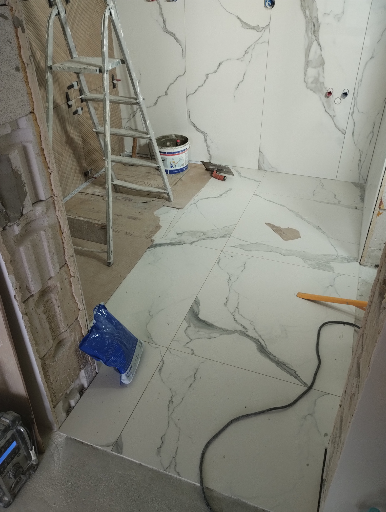 Remont łazienki w trakcie: białe płytki imitujące marmur na ścianach i podłodze, drabina, wiadro z klejem, narzędzia glazurnicze. Widoczne niedokończone fragmenty ścian.