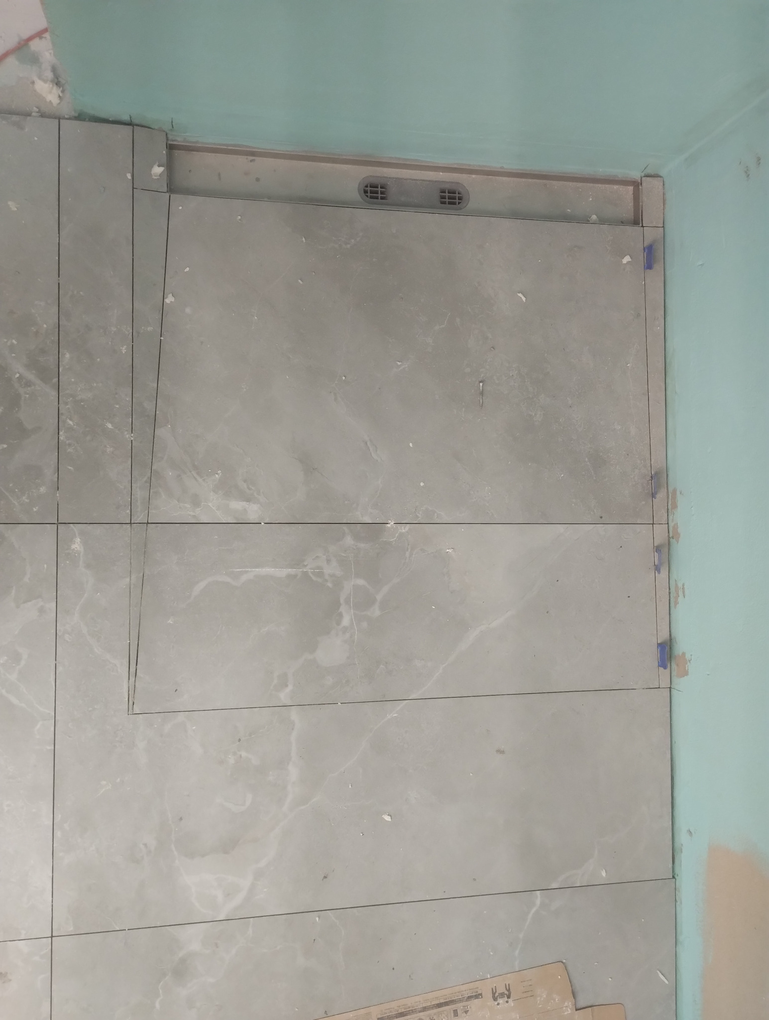 Ułożone szare płytki podłogowe z marmurkowym wzorem, widoczna kratka wentylacyjna przy ścianie, niebieska ściana w tle, etap prac wykończeniowych.