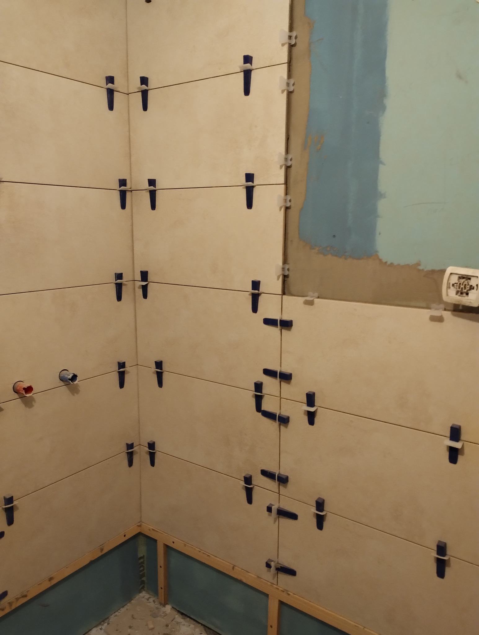 Ściana w trakcie układania płytek ceramicznych, z użyciem klipsów poziomujących. Widoczne fragmenty instalacji wodnej i przygotowane podłoże.