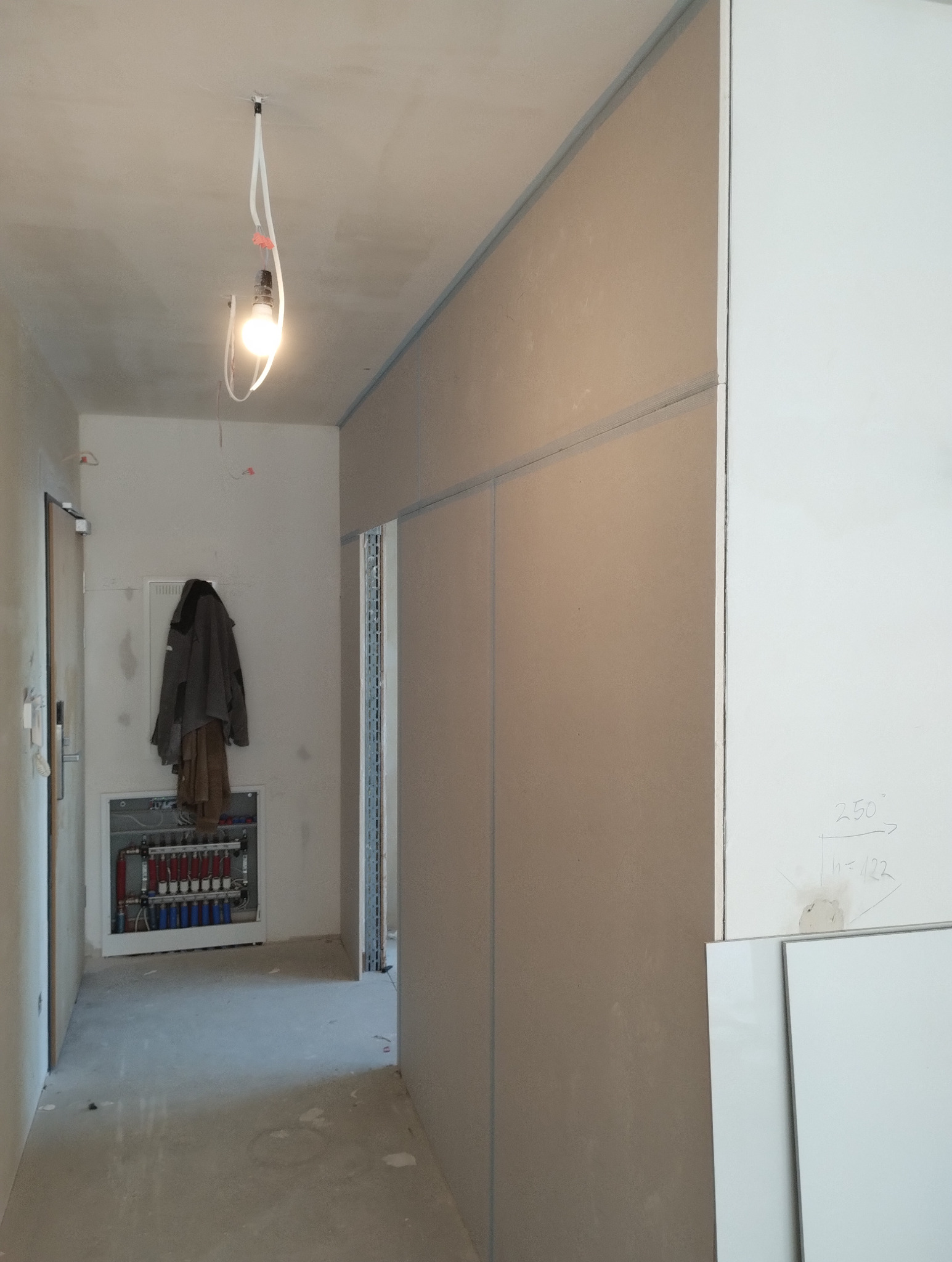 Korytarz w trakcie prac wykończeniowych: ściany z płyt kartonowo-gipsowych, widoczna instalacja grzewcza, surowy beton na podłodze, wisząca kurtka.