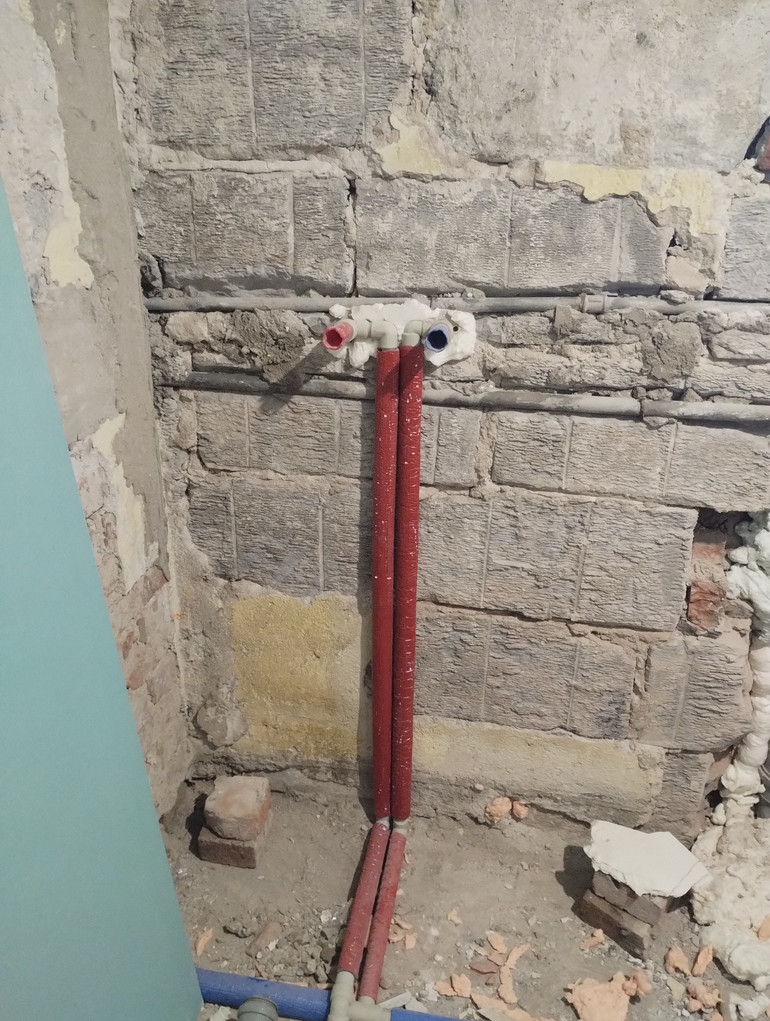 Czerwone rury grzewcze pionowo na tle surowej ściany z cegły i fragmentu płyty gipsowej. Widoczne łączenia i izolacja. Etap prac instalacyjnych w pomieszczeniu.