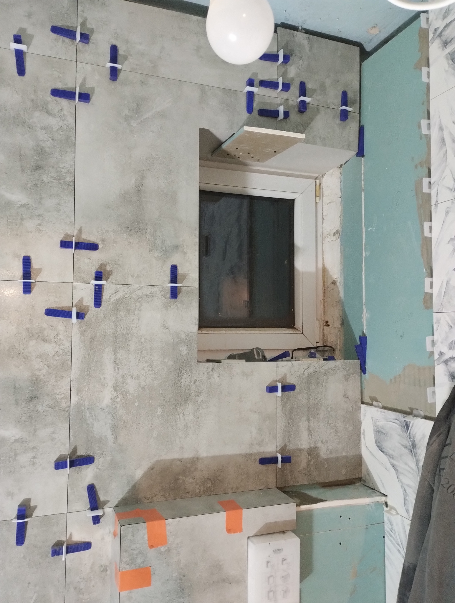 Ściana z szarych płytek w trakcie układania, z niebieskimi krzyżykami dystansowymi, widoczne okno i fragment instalacji. Prace glazurnicze w łazience.