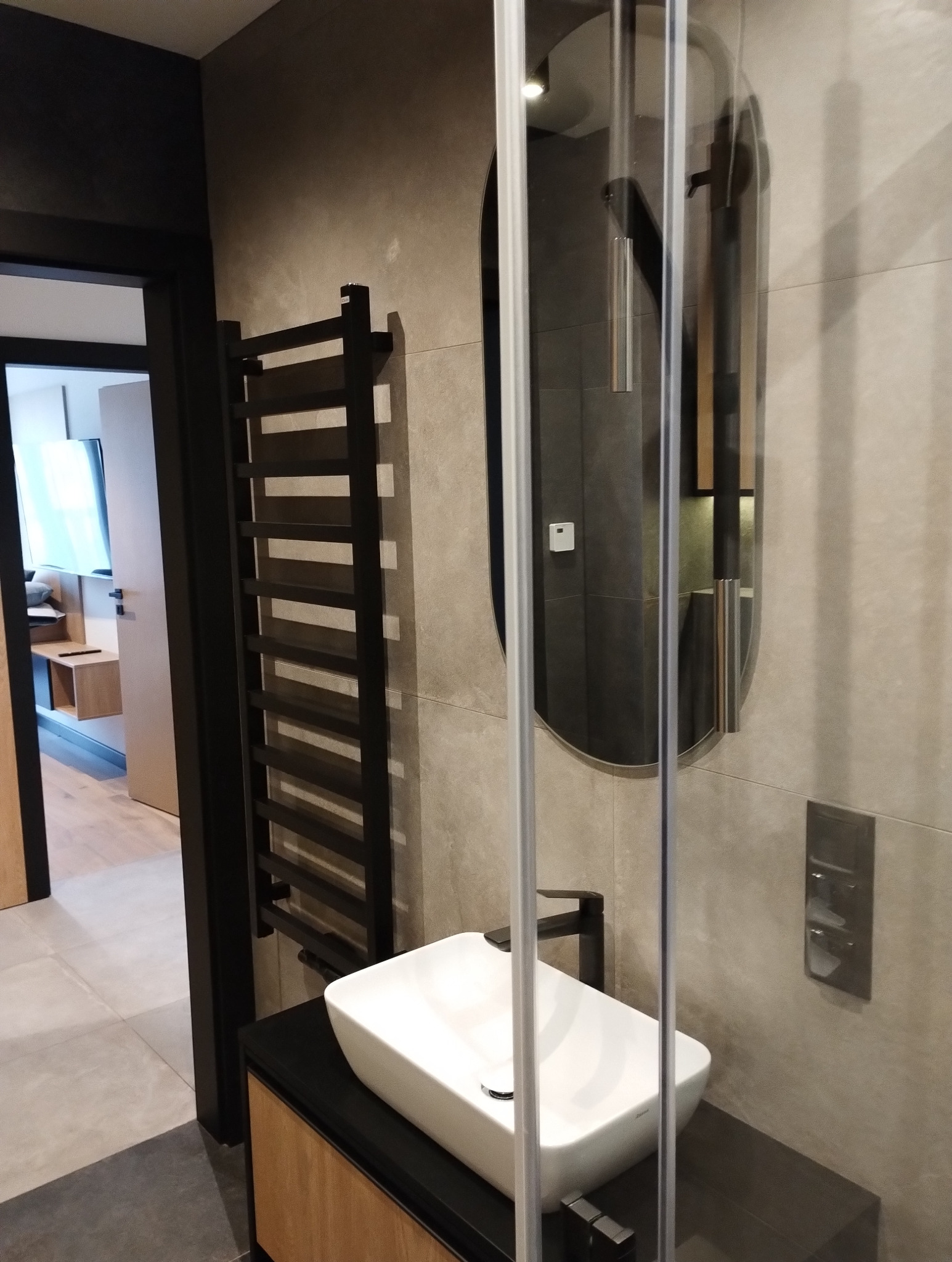 Nowoczesna łazienka z umywalką nablatową, czarną armaturą i grzejnikiem drabinkowym. Widoczna kabina prysznicowa i lustro w owalnej ramie. Stonowana kolorystyka.