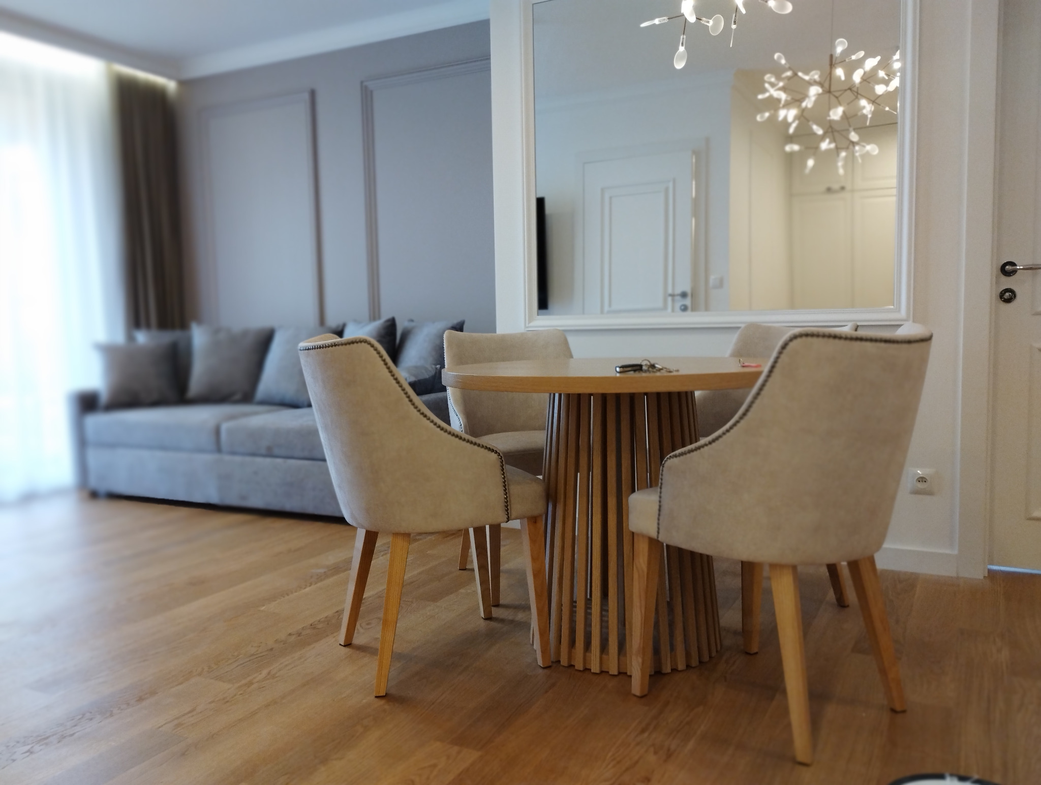 Jasne wnętrze: stół z krzesłami na drewnianej podłodze, w tle sofa i lustro odbijające nowoczesną lampę. Minimalistyczny design, stonowana kolorystyka.