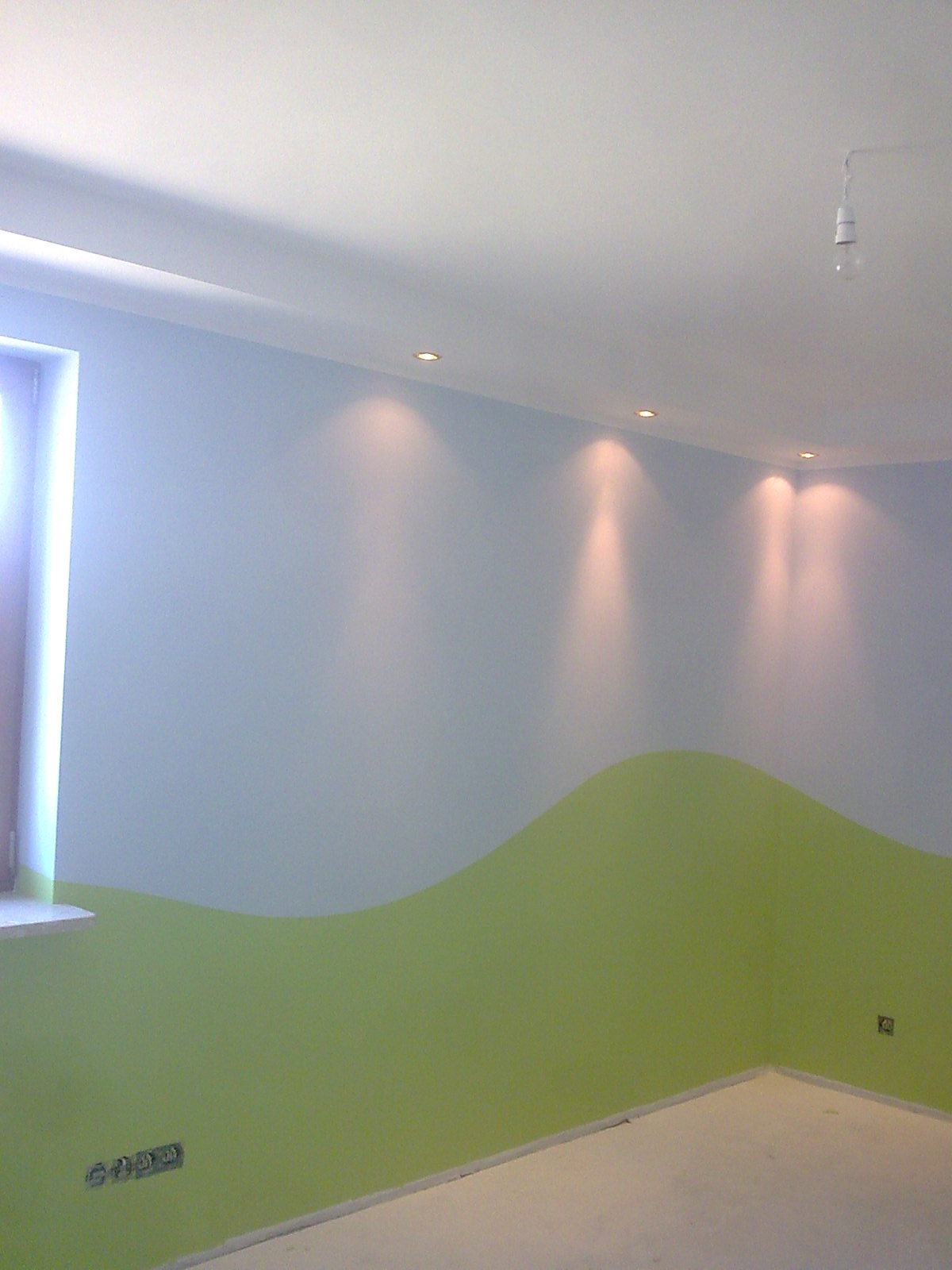Ściana w pokoju pomalowana na dwa kolory: górna część jasnoniebieska, dolna zielona w kształcie falującej linii, oświetlona punktowymi lampami w suficie.