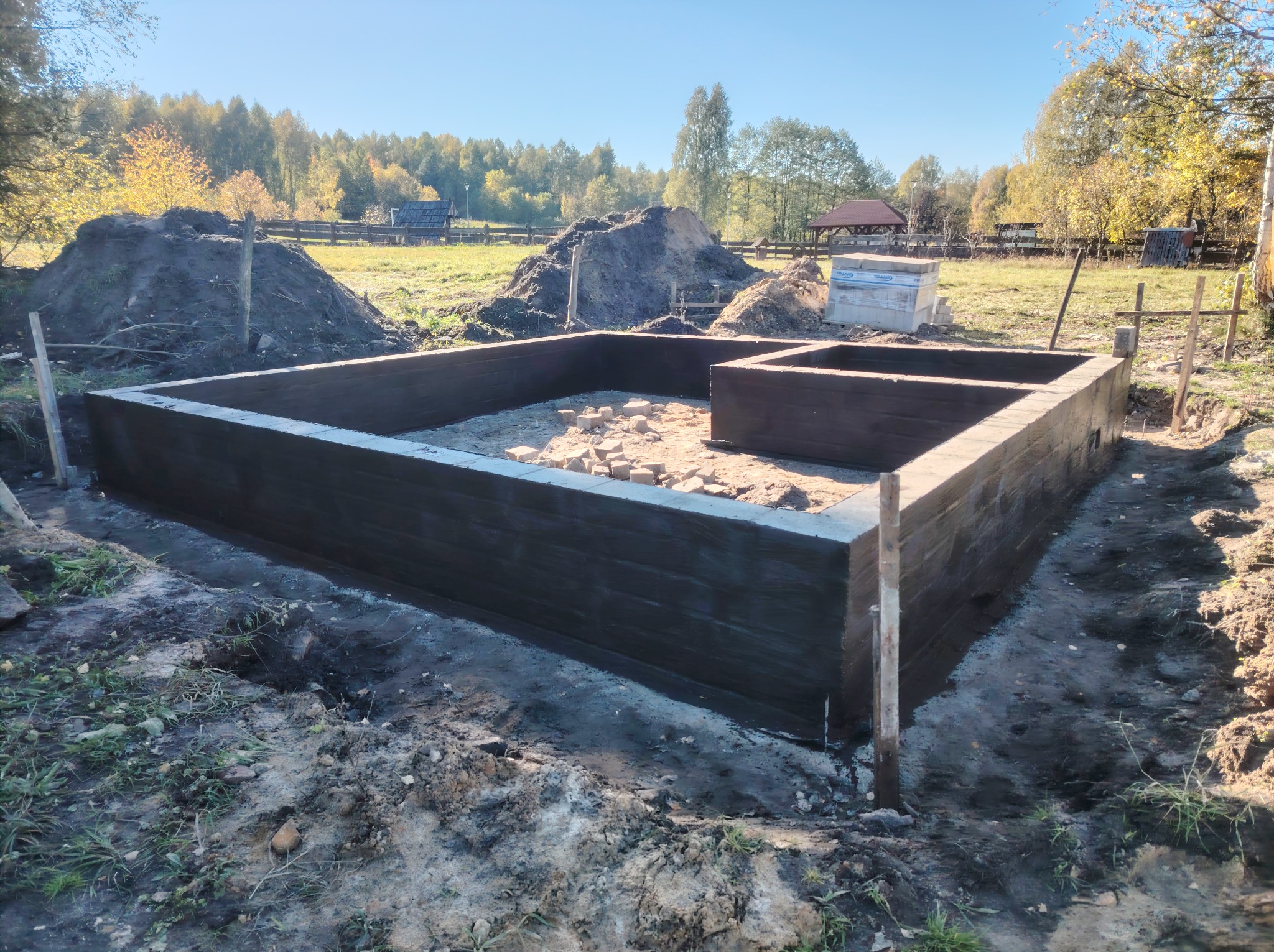 Zabezpieczone fundamenty pod budowę domu, widoczne cegły wewnątrz, ziemia i drzewa w tle, słoneczny dzień.