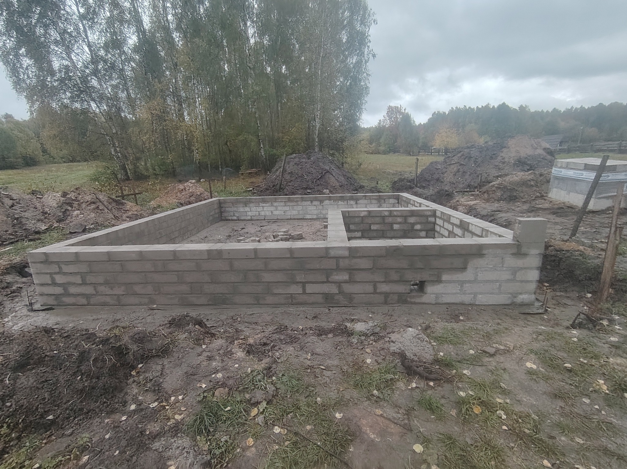 Fundamenty domu z bloczków betonowych na tle jesiennego krajobrazu. Widoczne wykopane ziemia i drzewa. Szary, pochmurny dzień.