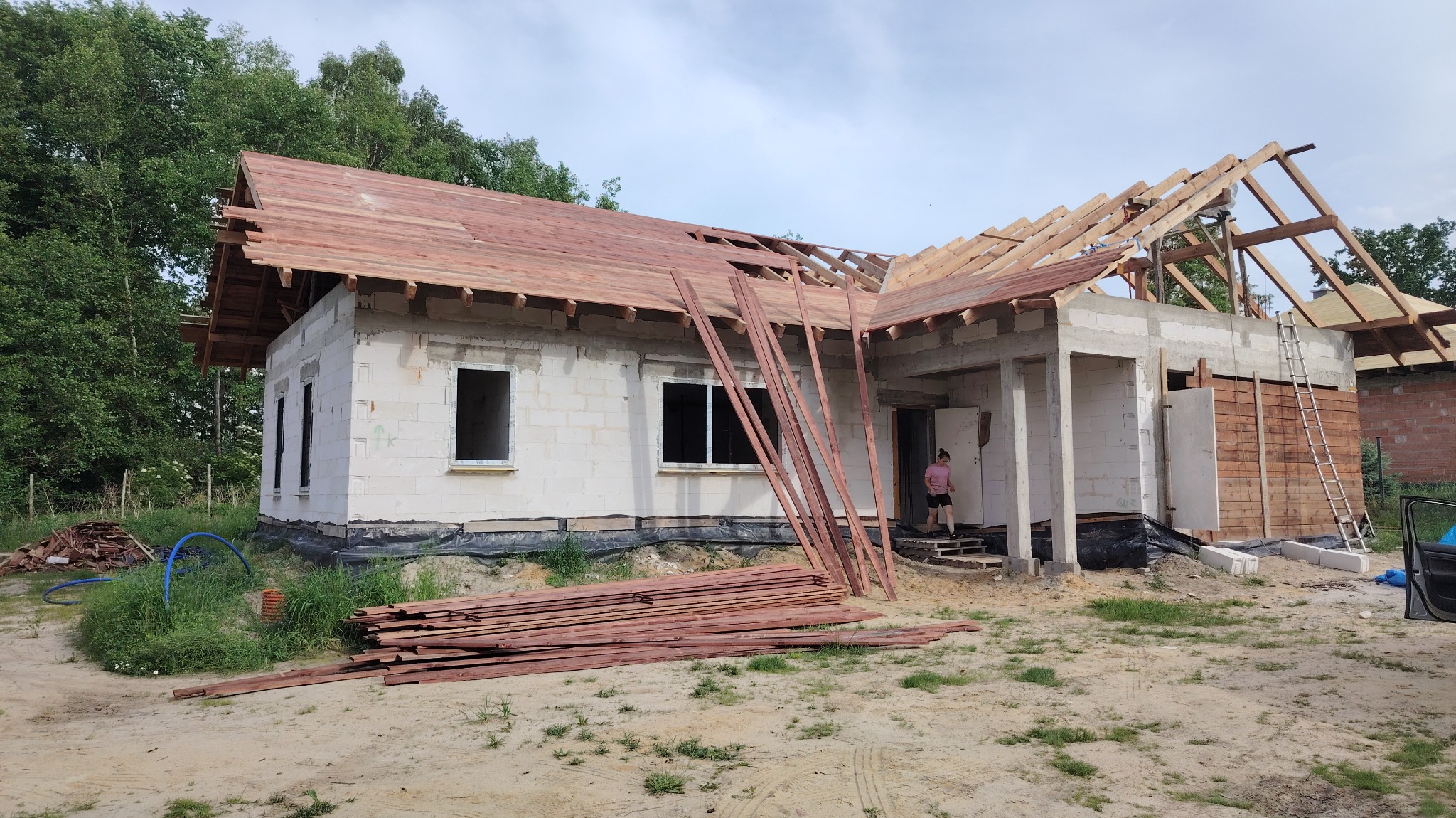 Budowa domu w trakcie: ściany z betonu komórkowego, częściowo pokryty dach z drewnianą konstrukcją. Na pierwszym planie stos desek. Osoba w tle.