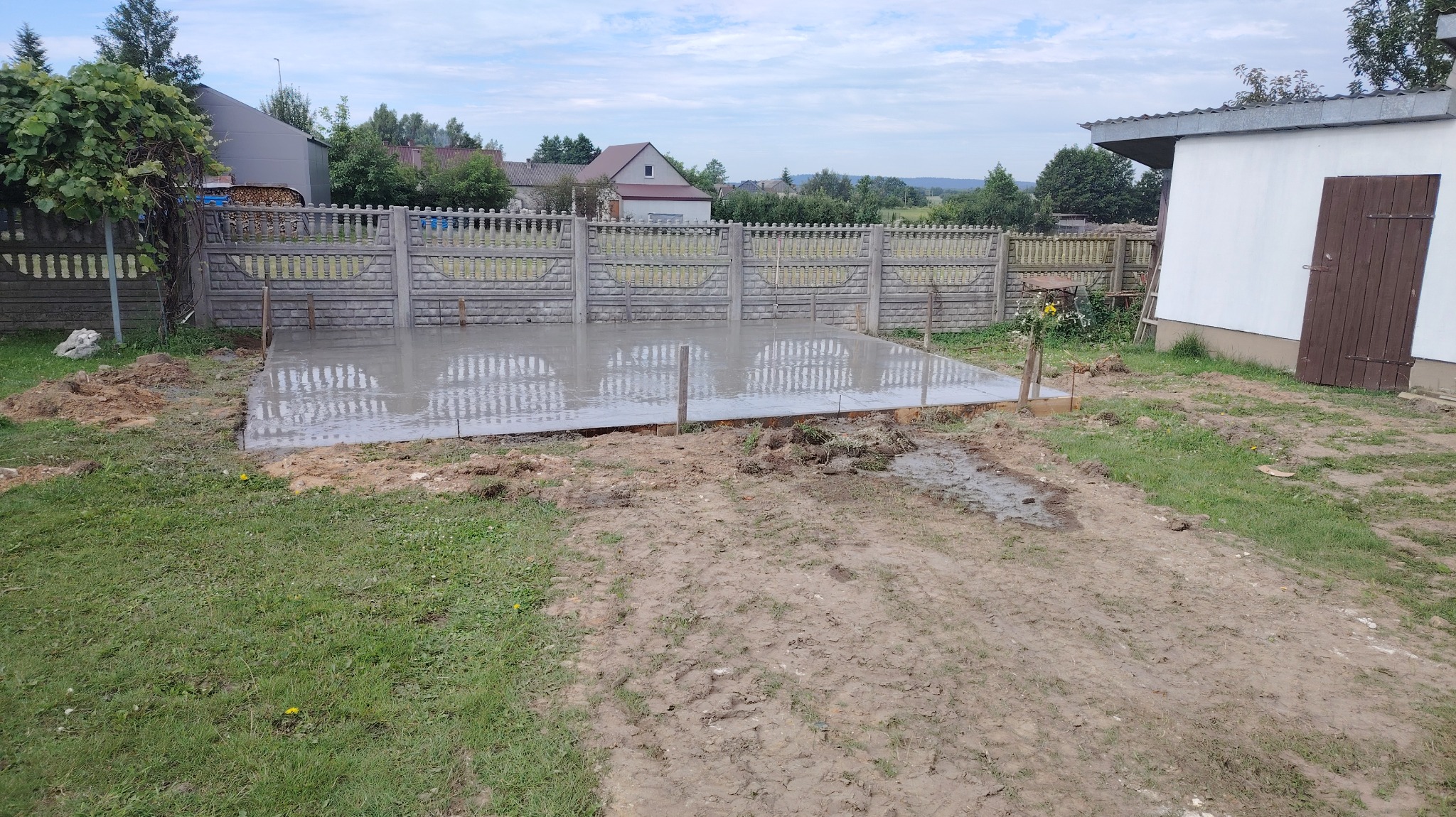 Wylany fundament pod budowę, widoczne drewniane szalunki i mokry beton. W tle ogrodzenie z prefabrykatów betonowych i budynek gospodarczy.