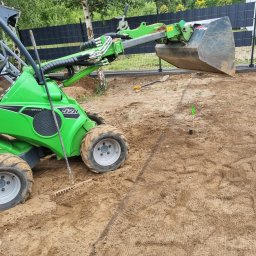 M&P Garden - Zielona miniładowarka Avant 528 na przygotowanym podłożu z siatką, prawdopodobnie pod trawnik. W tle czarne ogrodzenie panelowe i elementy placu zabaw.