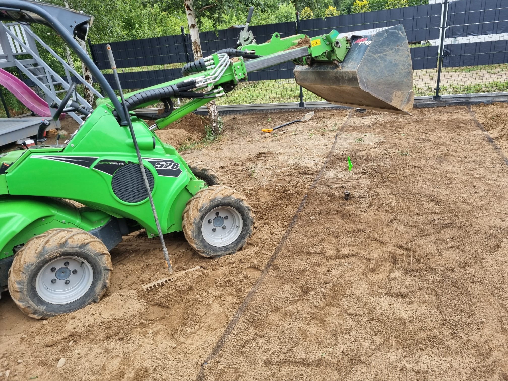 Zielona miniładowarka Avant 528 na przygotowanym podłożu z siatką, prawdopodobnie pod trawnik. W tle czarne ogrodzenie panelowe i elementy placu zabaw.