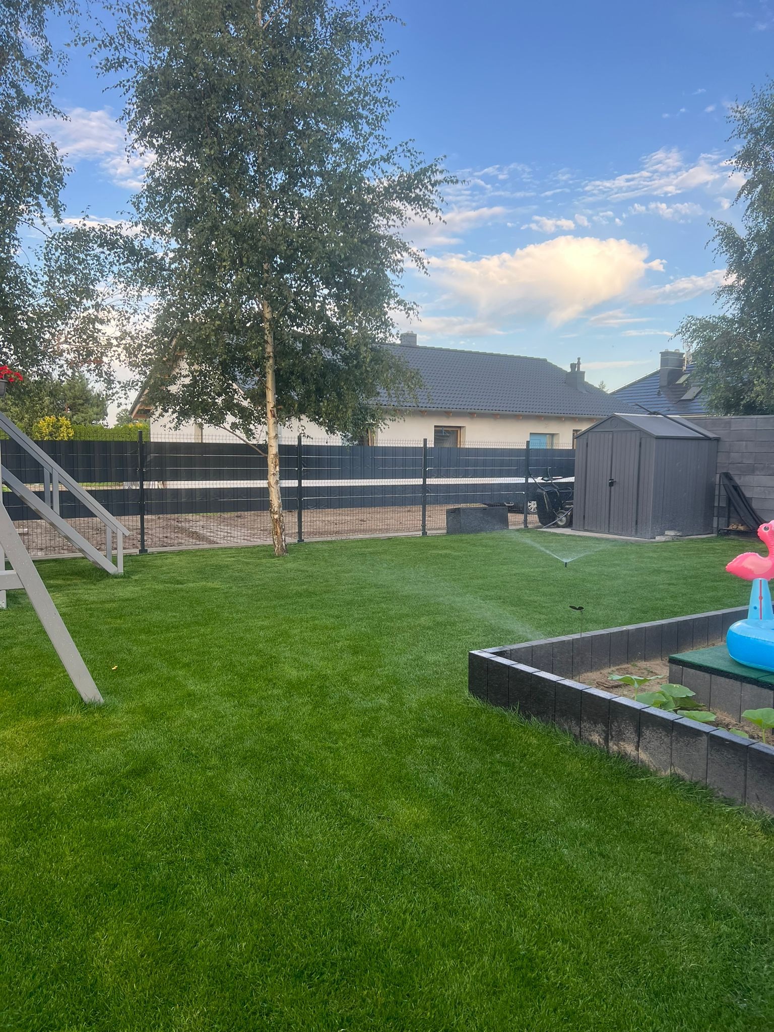 Zadbany trawnik z systemem nawadniania, ogrodzeniem panelowym i elementami dekoracyjnymi, takimi jak flaming, w tle dom z szarą dachówką i szopa.