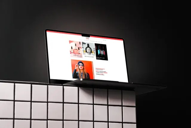 Laptop wyświetla stronę internetową z produktami perfumeryjnymi na tle czarnej ściany i białej, kafelkowej powierzchni. Widoczny ekran z ofertą sklepu internetowego.