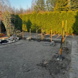 Lej&Tnij - Przygotowany teren pod trawnik z rolki: wyrównana ziemia, wbite słupki, narzędzia ogrodnicze i wał leżą na ziemi. W tle żywopłot z tui i drzewa.