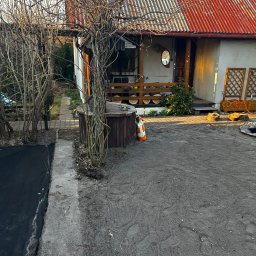 Lej&Tnij - Przygotowany teren pod ogród z widocznym domem z czerwonym dachem, drzewami bez liści i workiem nawozu. Czarna agrowłóknina częściowo rozłożona.