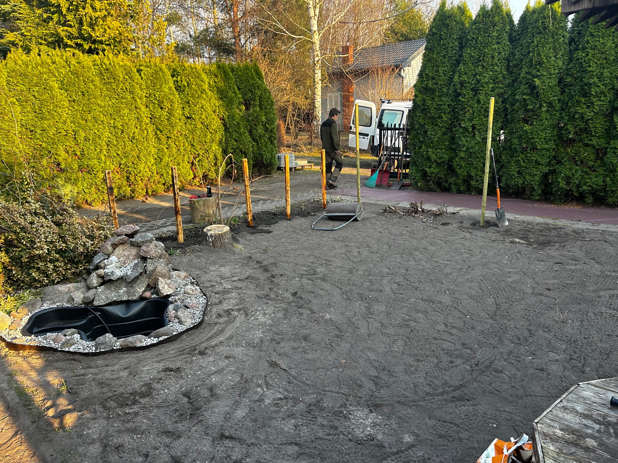Etap budowy oczka wodnego w ogrodzie: wykopany dół z folią, obłożony kamieniami, widoczne narzędzia i pracownik w tle. Przygotowany teren pod dalsze prace.