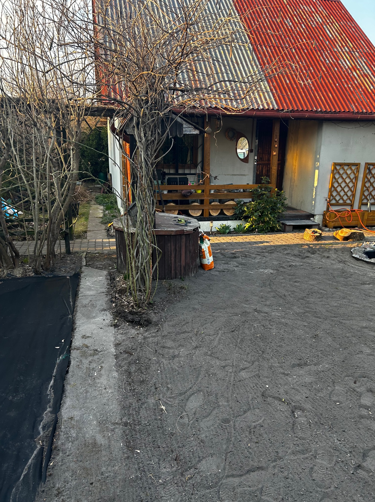Przygotowany teren pod ogród z widocznym domem z czerwonym dachem, drzewami bez liści i workiem nawozu. Czarna agrowłóknina częściowo rozłożona.