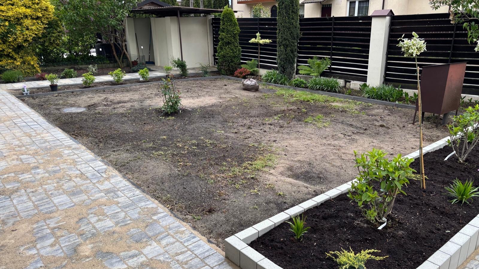 Przygotowany teren pod trawnik z obrzeżami i nasadzeniami wzdłuż ogrodzenia. Widoczny fragment chodnika z kostki brukowej i pojedyncze rośliny.