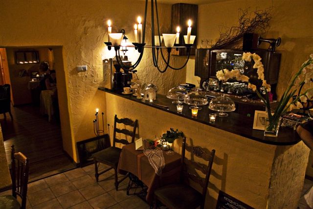 Urokliwe wnętrze herbaciarni w stylu retro z ciemnym barem, dekoracyjnym oświetleniem świecowym, stolikiem nakrytym do posiłku i ekspozycją deserów pod szklanymi kloszami.