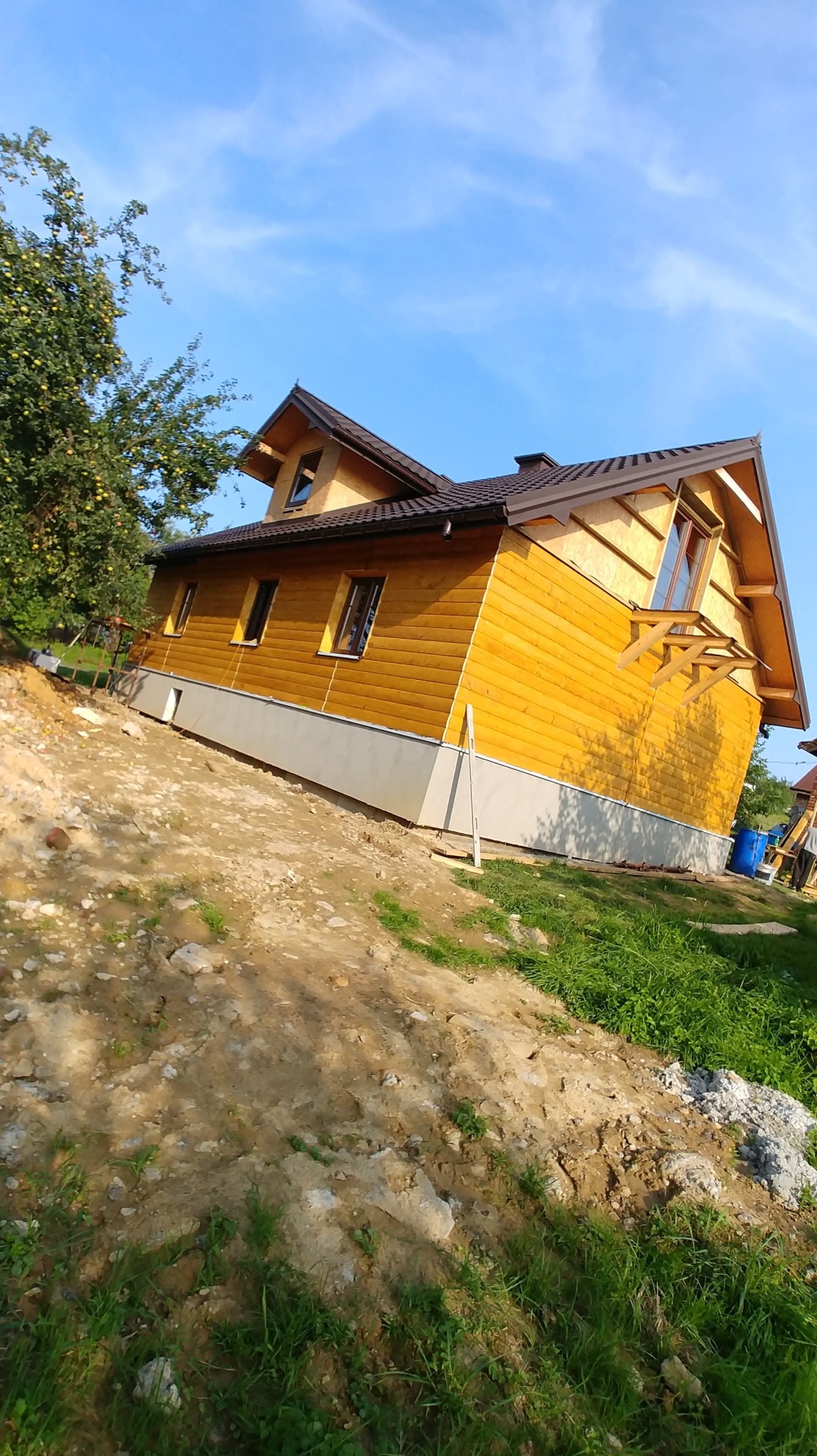 Żółty dom z drewnianą elewacją i brązowym dachem w trakcie budowy, widoczny z dołu pod kątem. W tle błękitne niebo i zieleń drzew.