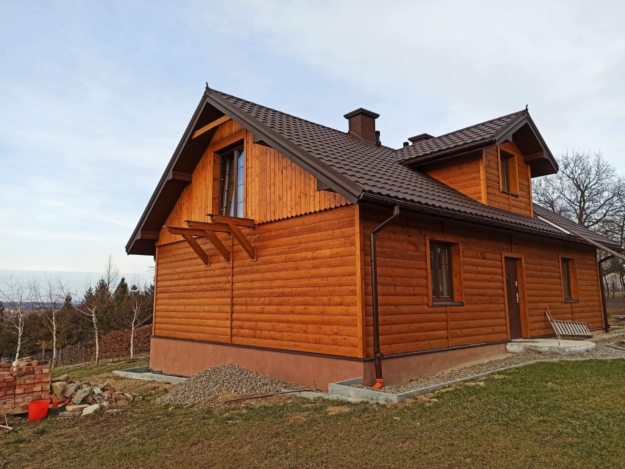 Drewniany dom z brązowym dachem w Gorlicach, widok z zewnątrz. Widoczne detale wykończenia, stos cegieł i kamieni obok budynku. Ujęcie z dołu, pochmurne niebo.