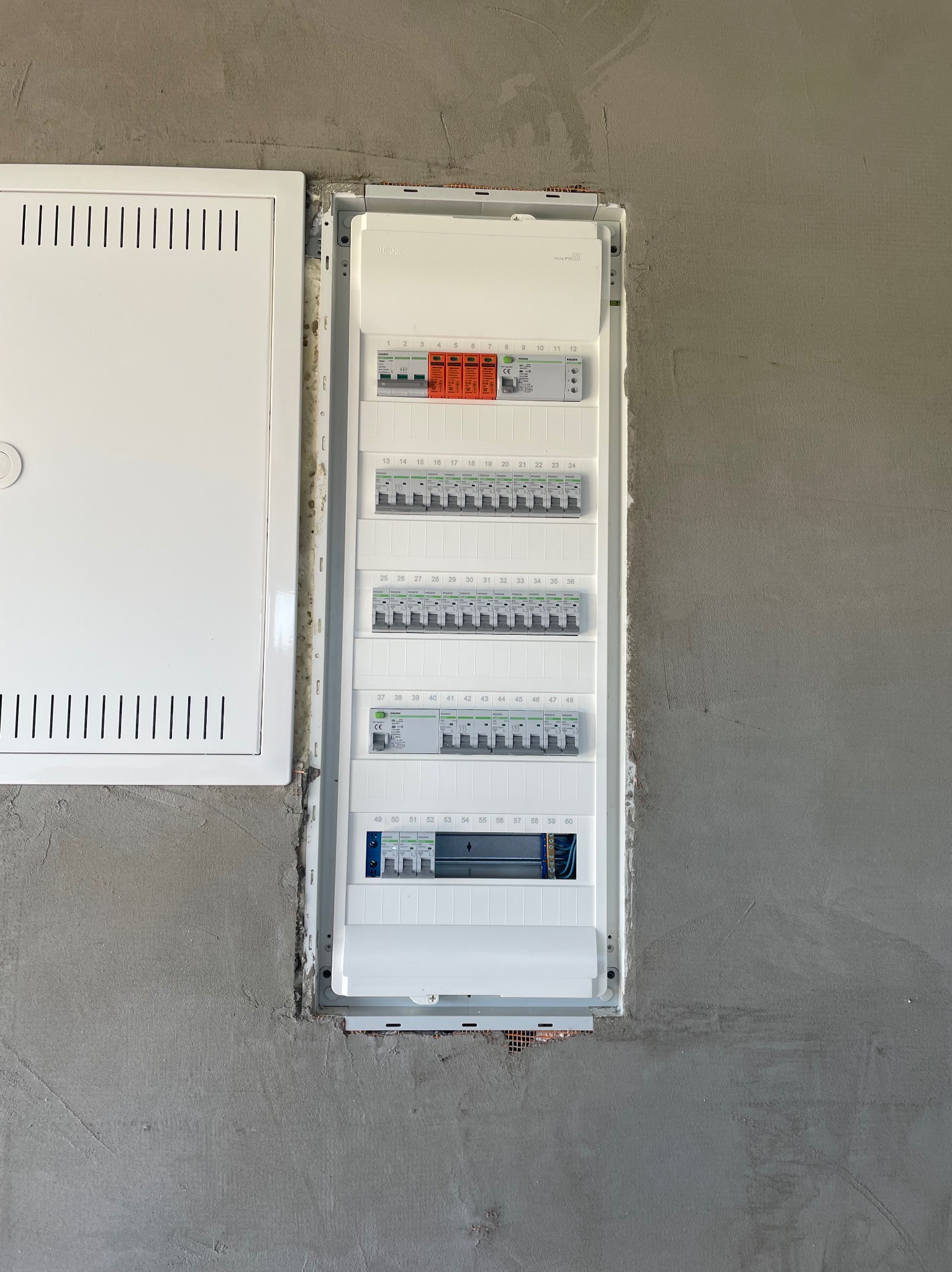 Rozdzielnica elektryczna Hager w surowej ścianie, widoczne zabezpieczenia i numeracja obwodów. Obok puszka instalacyjna z wentylacją. Szary beton w tle.
