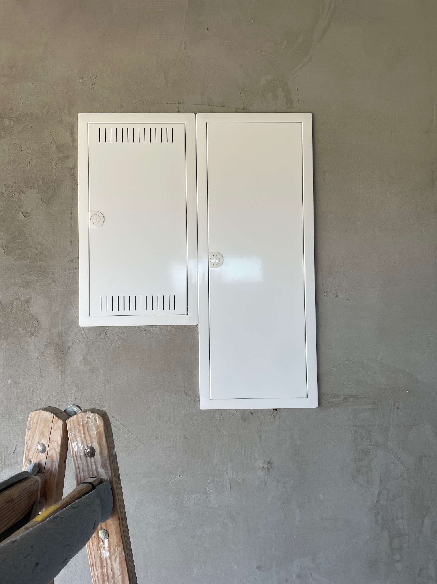 Dwie białe skrzynki elektryczne zamontowane na szarej, surowej ścianie. Po lewej skrzynka z wentylacją, po prawej podłużna. Fragment drabiny malarskiej w lewym dolnym rogu.