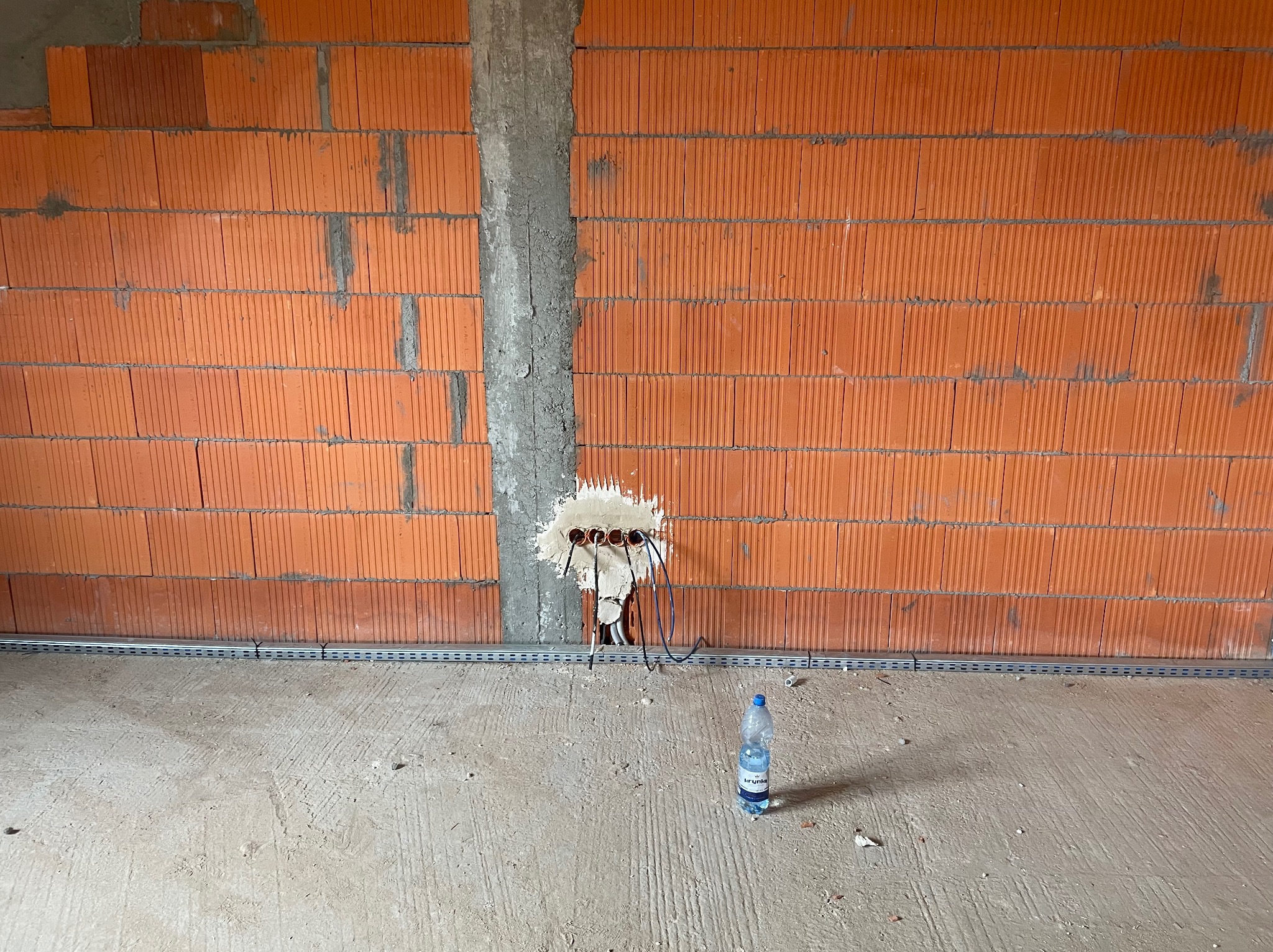 Ceglana ściana z puszkami elektrycznymi i wystającymi przewodami, zamontowane koryto kablowe. W tle surowy beton i butelka wody mineralnej.