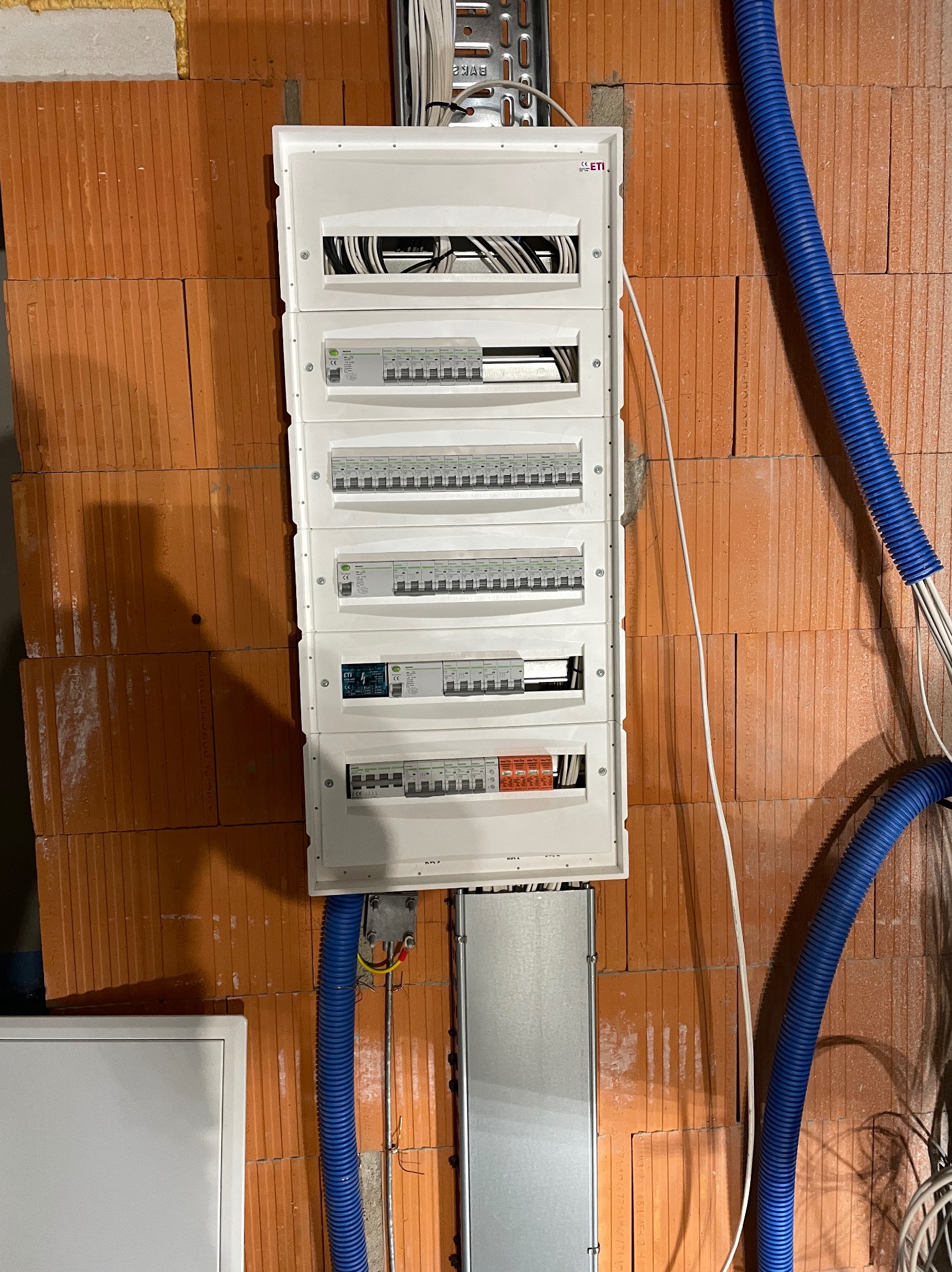 Rozdzielnica elektryczna z zabezpieczeniami na ceglanej ścianie, widoczne kable w peszlach. Instalacja w trakcie montażu, surowy stan.