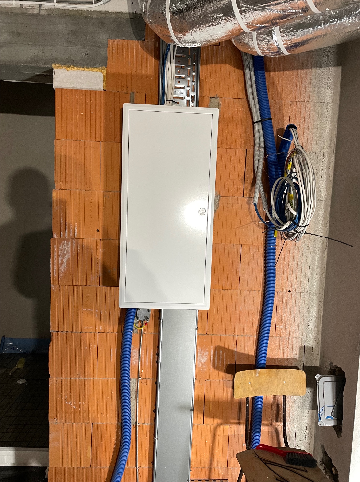 Biała skrzynka elektryczna na ceglanej ścianie z niebieskimi rurami instalacyjnymi i przewodami. Widoczne elementy instalacji wentylacyjnej pod sufitem.