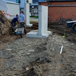 Marcin Bieniek Ogrody-Bieniek - Żółta koparka umieszcza betonowe elementy fundamentu. Dwóch pracowników budowlanych przy taczce. Widoczne narzędzia i przygotowany teren pod budowę.