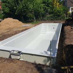 Marcin Bieniek Ogrody-Bieniek - Montaż gotowego basenu z tworzywa w wykopie, widoczne narzędzia i hałda ziemi. Słoneczny dzień, zielone drzewa w tle. Etap instalacji.