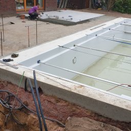 Marcin Bieniek Ogrody-Bieniek - Budowa basenu: wykop, betonowa obudowa, instalacja. W tle pracownik układa płyty chodnikowe. Widoczne narzędzia i materiały budowlane.