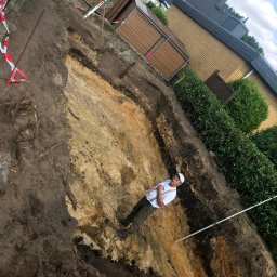 Marcin Bieniek Ogrody-Bieniek - Wykop pod fundamenty domu jednorodzinnego z widocznym pracownikiem z łopatą, zabezpieczony taśmami ostrzegawczymi, w tle zabudowa mieszkalna.