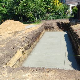 Marcin Bieniek Ogrody-Bieniek - Wykop pod fundament z wylaną betonową płytą w ogrodzie. Widoczne zabezpieczenia taśmą ostrzegawczą i sterta ziemi. Prace ziemne w toku, przygotowanie pod budowę.