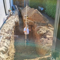 Marcin Bieniek Ogrody-Bieniek - Wykop pod fundamenty domu jednorodzinnego, widok z kabiny koparki. Pracownik z łatą niwelacyjną kontroluje głębokość wykopu. W tle drewno na opał i zielony żywopłot.