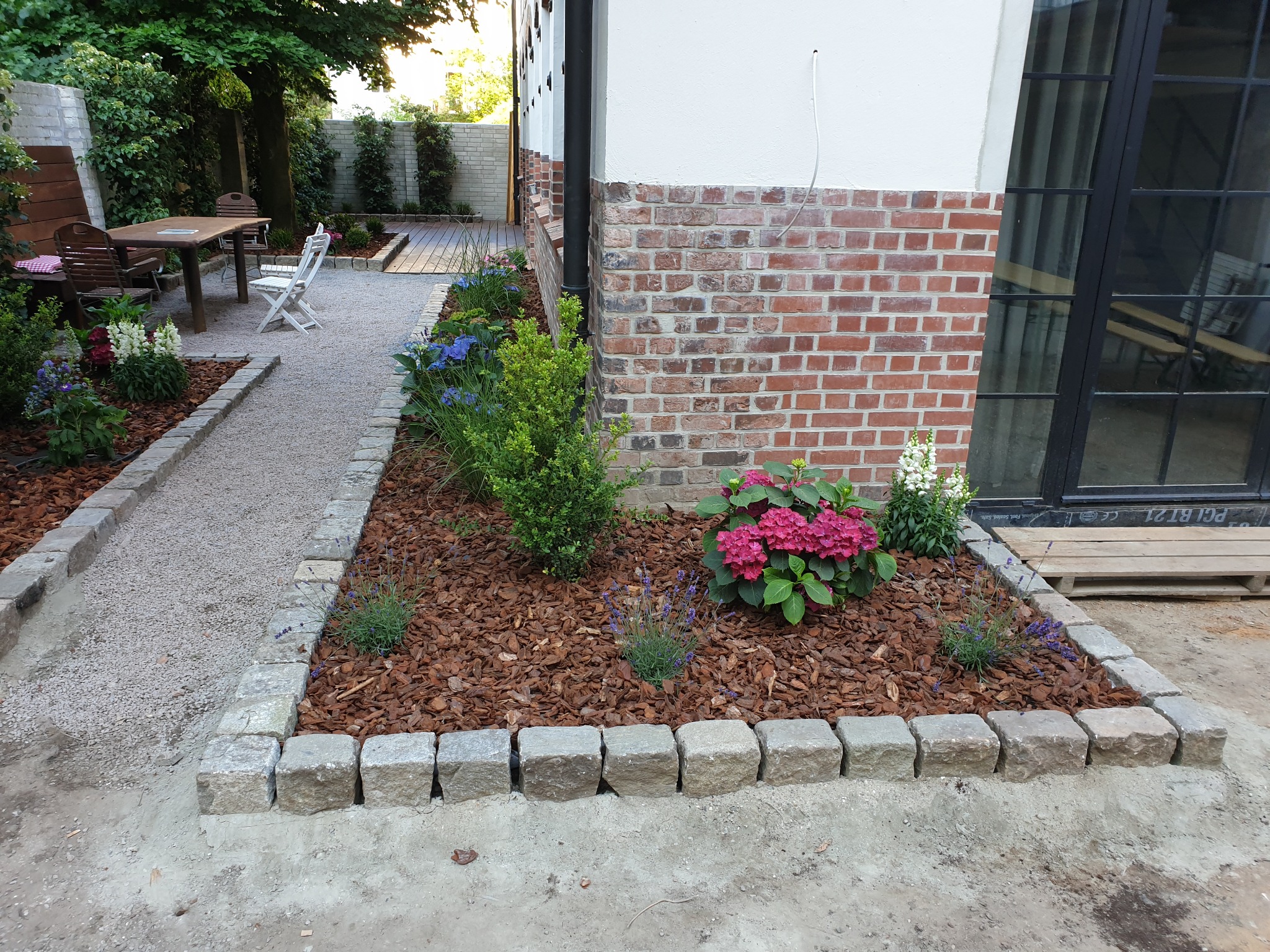 Kwadratowa rabata z obrzeżem z kostki granitowej, wypełniona korą i roślinami, w tle ścieżka z kamyczków i drewniany taras, widok na budynek z ceglaną elewacją.