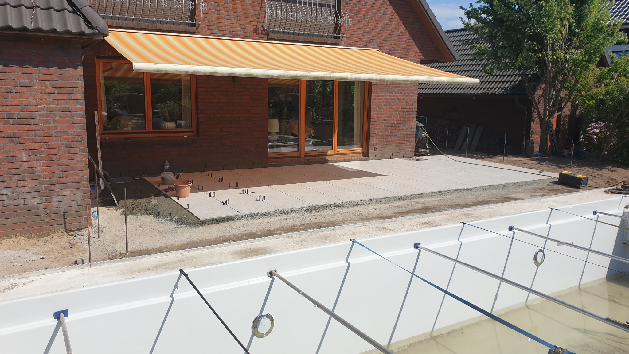 Montaż tarasu z jasnych płyt przy basenie, widoczne narzędzia i elementy mocujące. Częściowo zadaszony taras z markizą w paski przy ceglanym budynku.