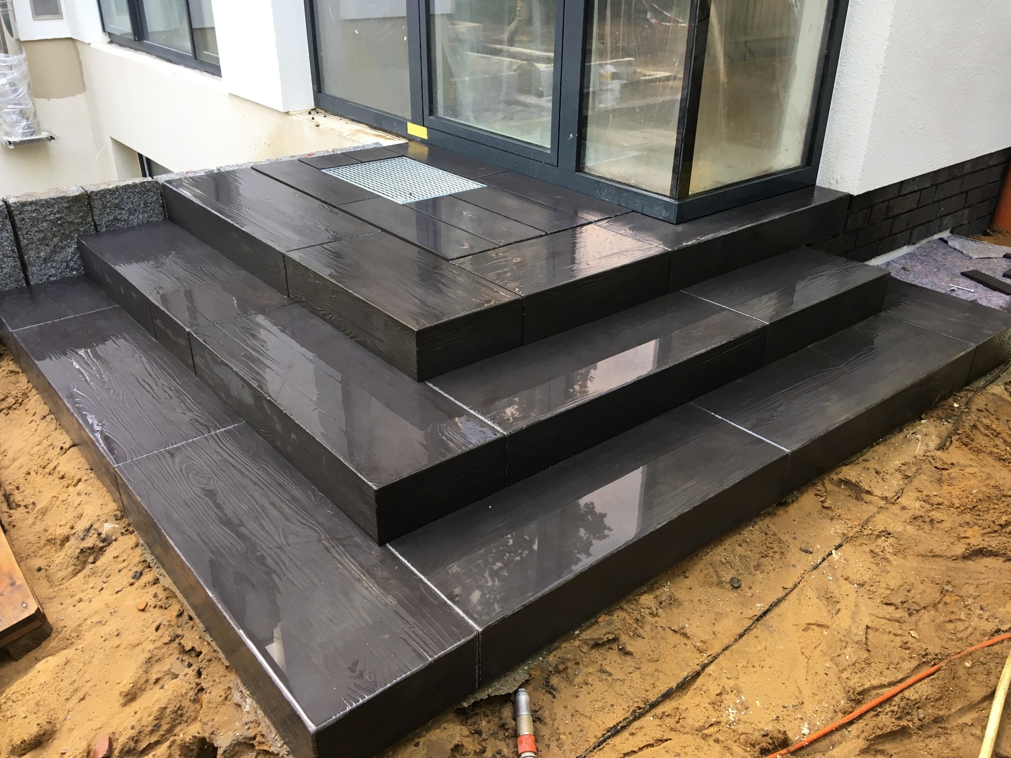 Nowoczesne schody tarasowe z ciemnych płyt imitujących drewno, prowadzące do przeszklonych drzwi. Widoczne elementy wykończeniowe i przygotowany teren.