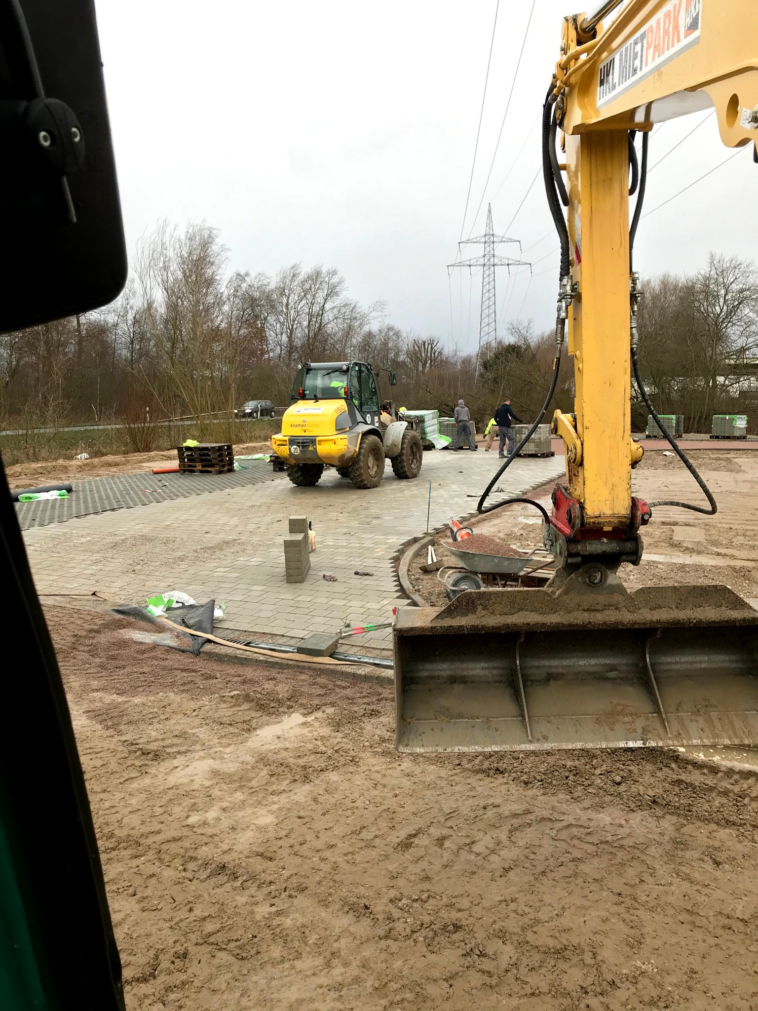 Żółta koparka i miniładowarka na placu budowy z częściowo ułożoną kostką. W tle słupy wysokiego napięcia i pracownicy. Widok z kabiny maszyny.