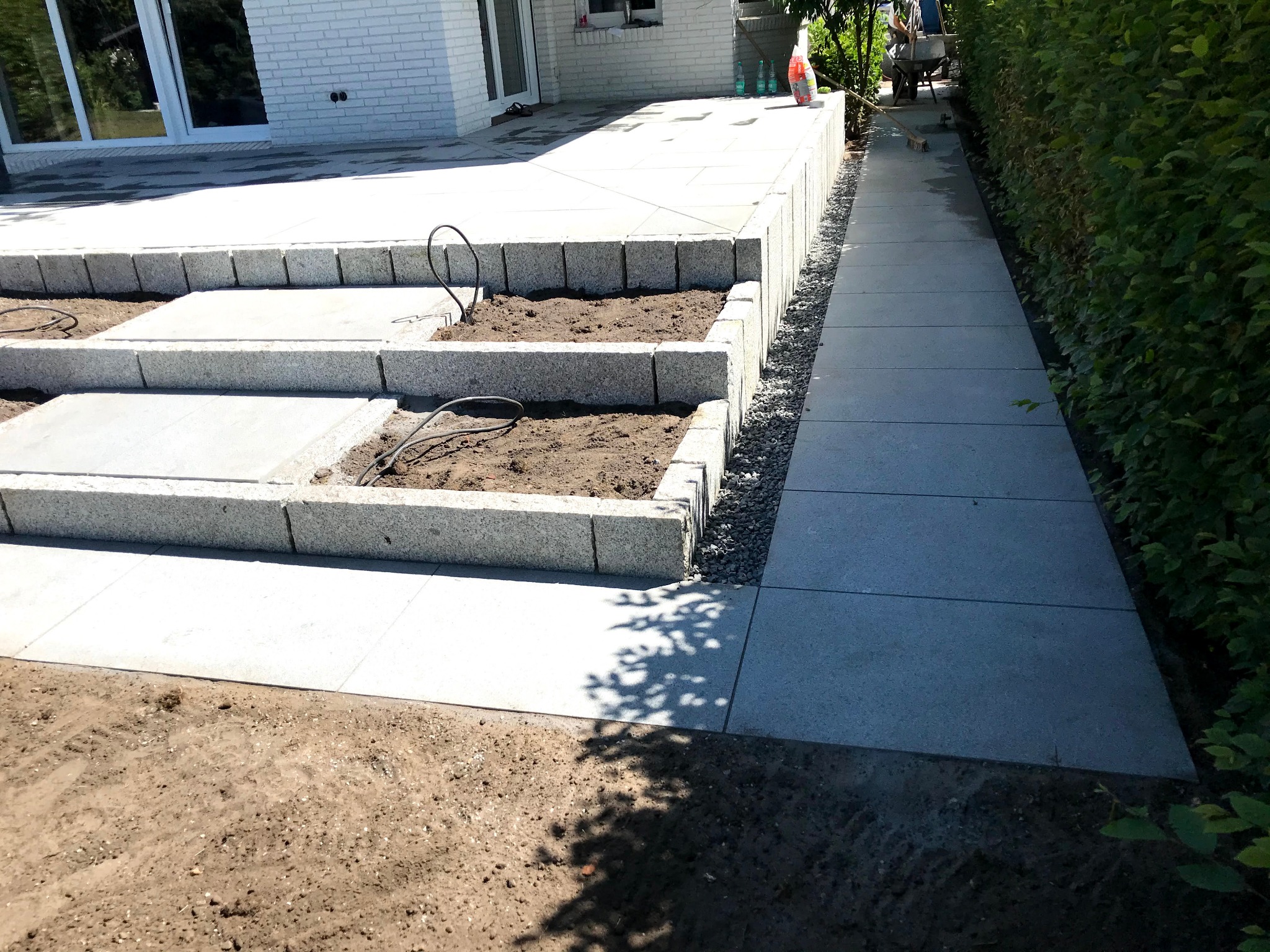 Jasnoszara, kamienna ścieżka z obrzeżami i schodami prowadząca wzdłuż żywopłotu. Widoczne przygotowane rabaty z ziemią. Ujęcie z dołu, w tle budynek.