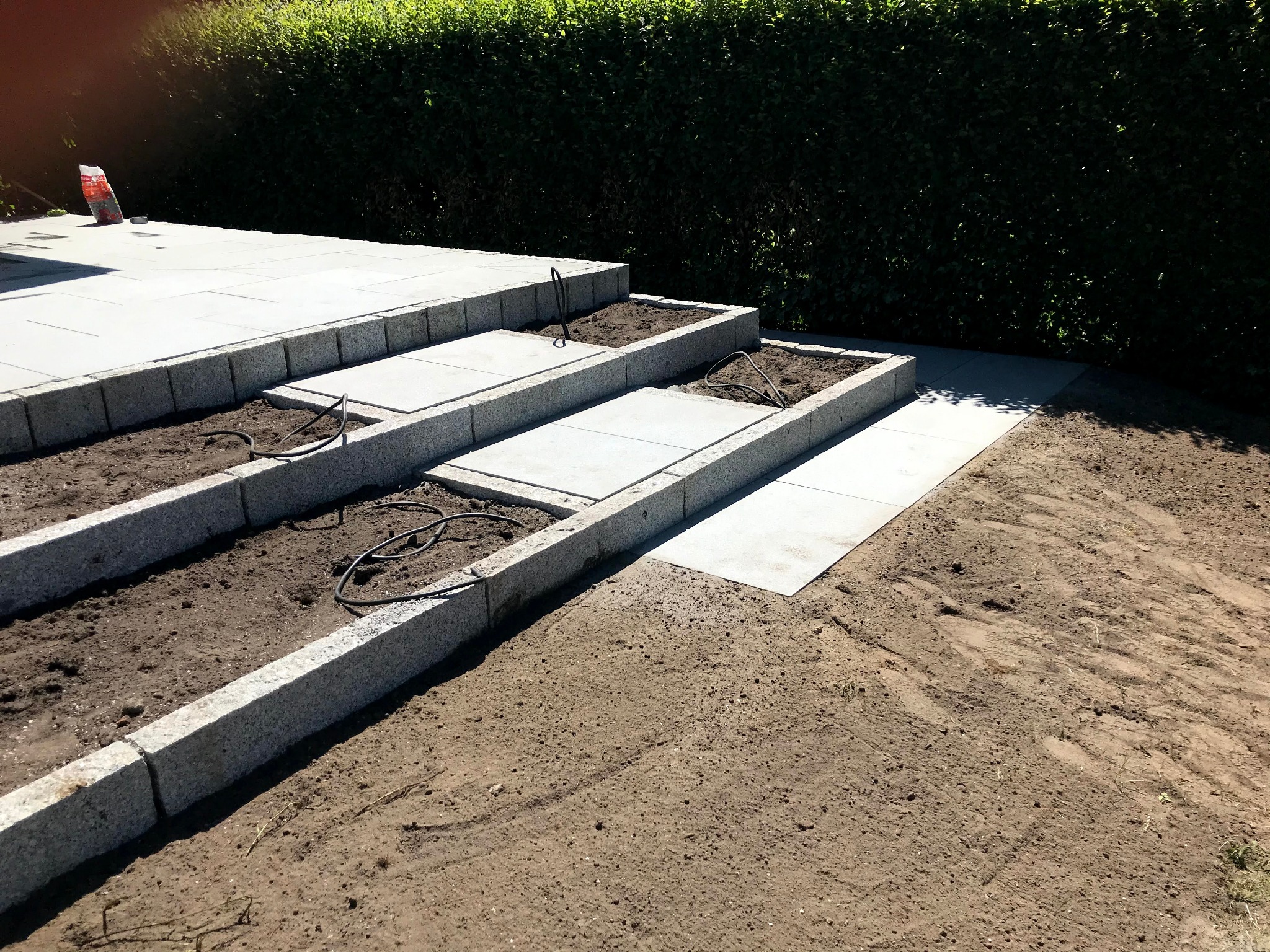 Schodkowy taras z kostki granitowej w ogrodzie, z przygotowanymi rabatami ziemnymi i instalacją nawadniającą. Tło stanowi zielony żywopłot.