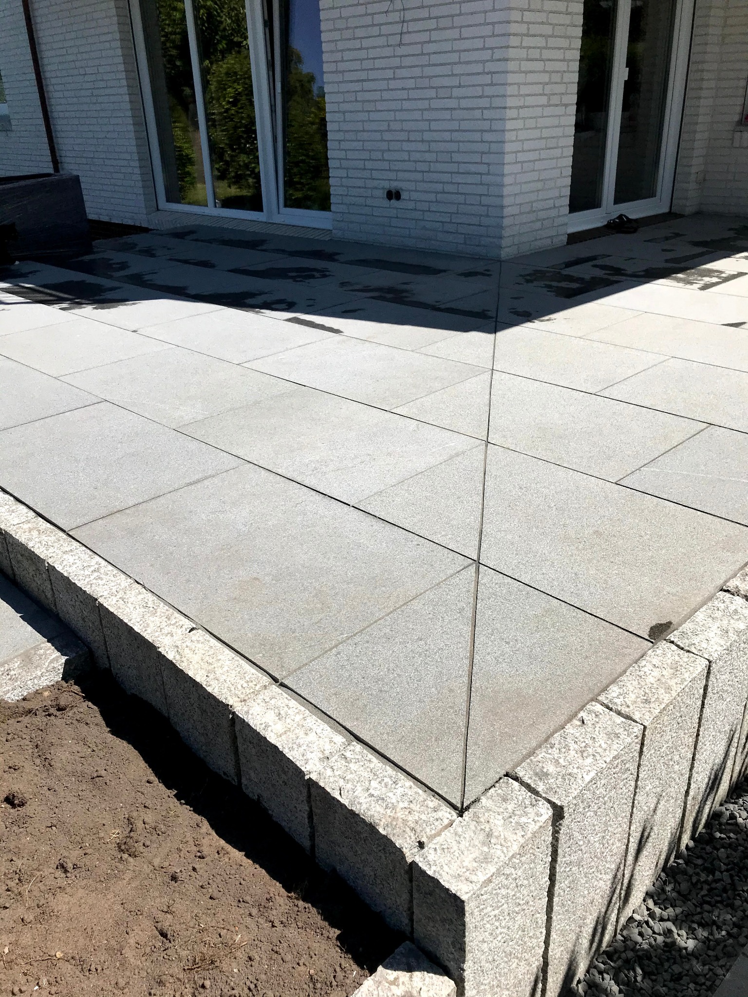 Jasnoszary taras z kamiennych płyt, obrzeża z kostki granitowej, widok na biały budynek z dużymi oknami. Częściowo widoczna ziemia przygotowana pod nasadzenia.