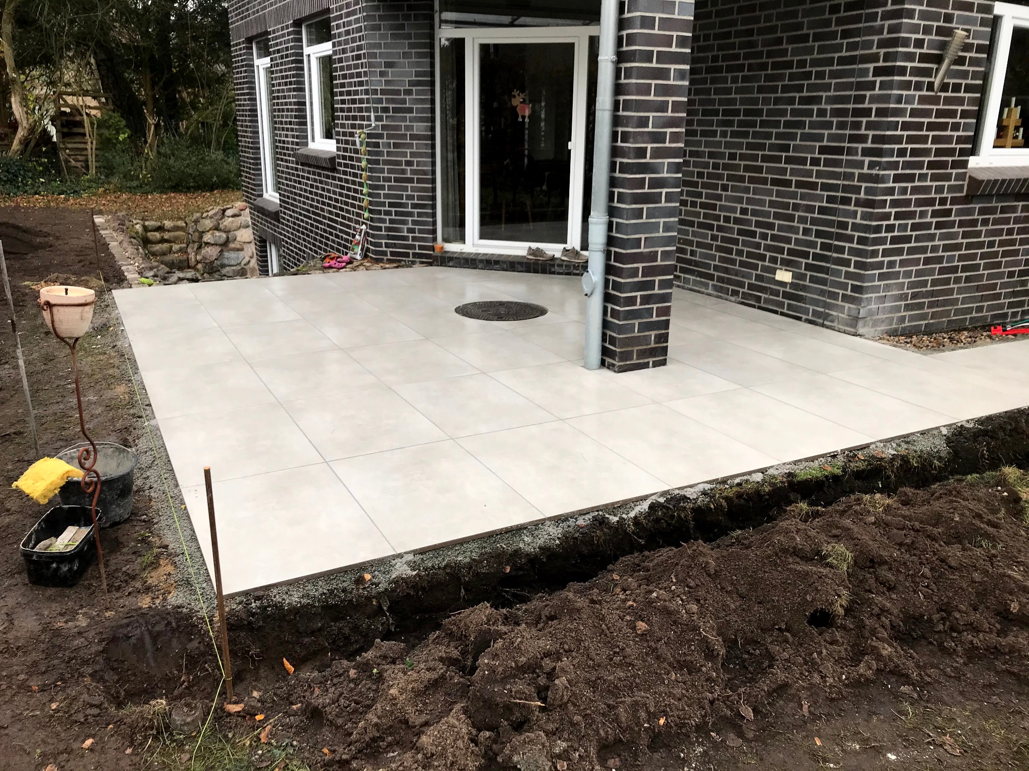 Jasne, kwadratowe płyty tarasowe ułożone przy ciemnej, ceglanej ścianie domu. Widoczne narzędzia i wykopana ziemia sugerują trwające prace wykończeniowe.