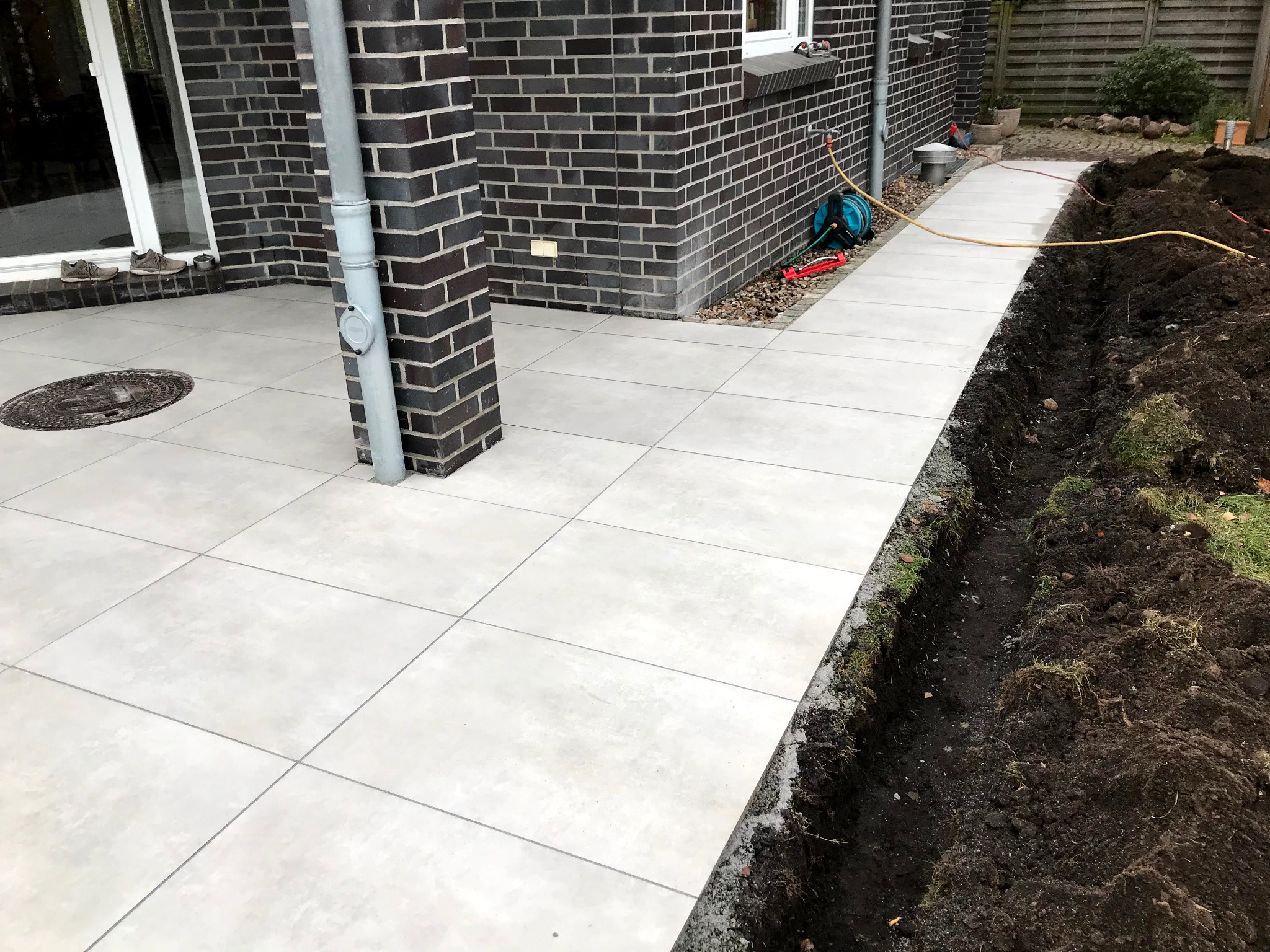 Jasnoszara, kwadratowa nawierzchnia z płytek przy ciemnej ceglanej ścianie domu. W tle widoczny wykop pod obrzeże. Częściowo ułożony chodnik.