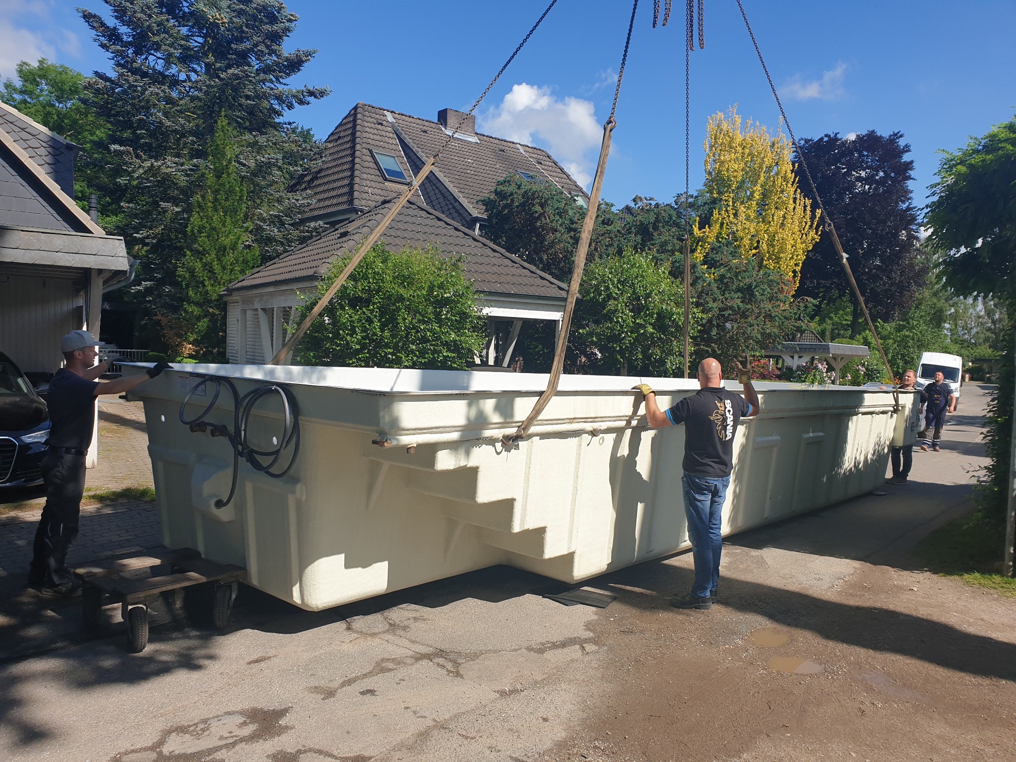 Montaż gotowego basenu ogrodowego z pomocą dźwigu. Widoczni pracownicy w trakcie instalacji. Tło: dom jednorodzinny i zieleń.