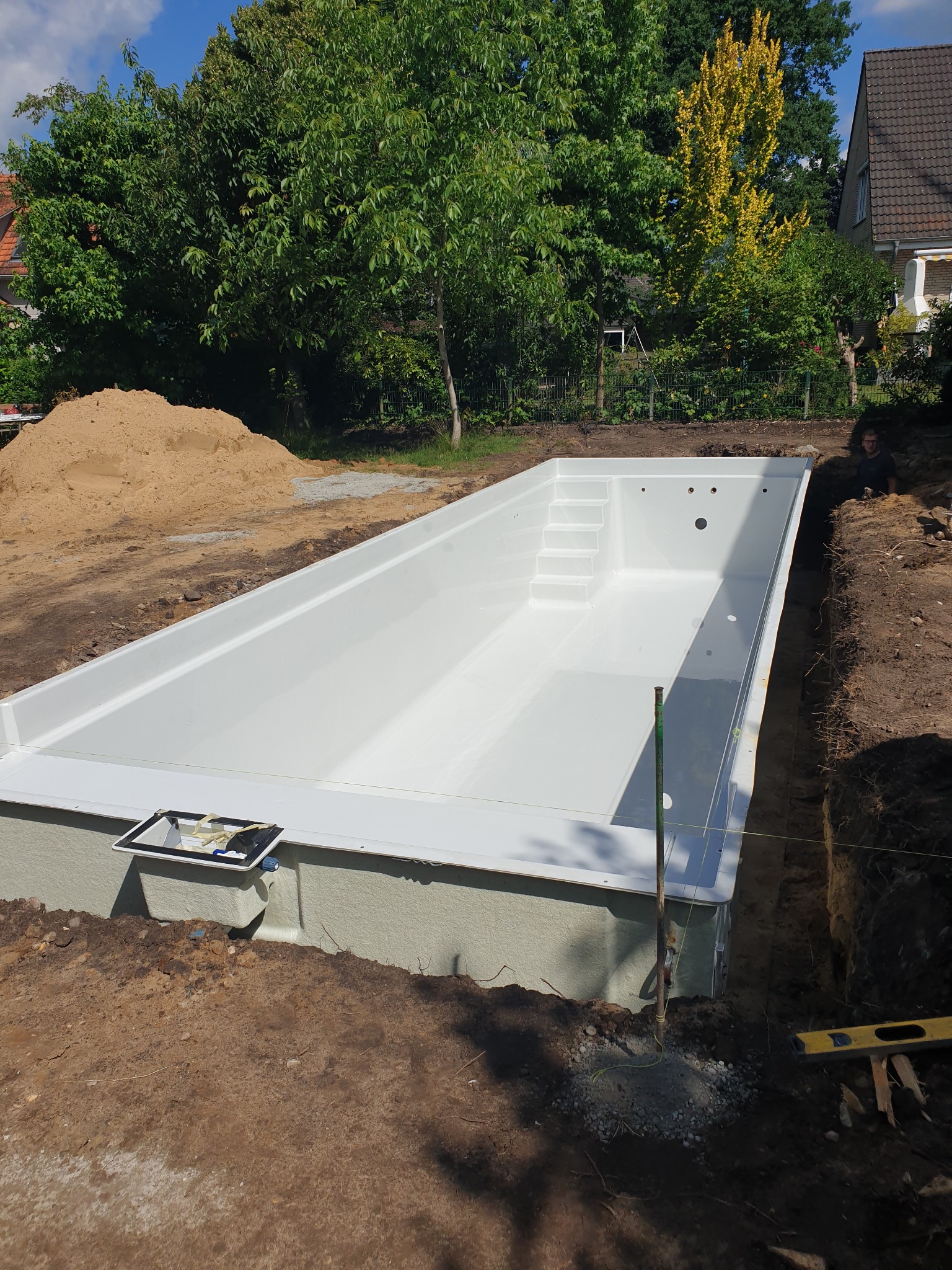 Montaż gotowego basenu z tworzywa w wykopie, widoczne narzędzia i hałda ziemi. Słoneczny dzień, zielone drzewa w tle. Etap instalacji.